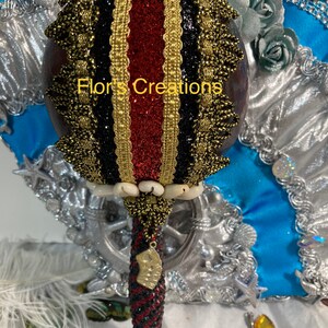 Peut inclure: Un hochet rouge, noir et or avec un manche perlé et un charme en or. Le hochet est décoré de paillettes et de dentelle. Le texte "Flor's Creations" est visible sur le hochet.