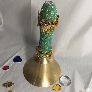Peut inclure: Une cloche dorée avec une poignée recouverte de strass turquoise. La poignée est décorée de breloques dorées, dont un éléphant, une main et une petite couronne.