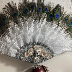 Peut inclure: Un &eacute;ventail en dentelle blanche avec un ornement argent&eacute; et blanc de fleurs et de strass. L'&eacute;ventail est entour&eacute; de plumes de paon.