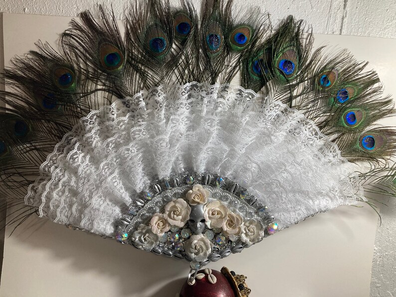 Peut inclure: Un &eacute;ventail en dentelle blanche avec un ornement argent&eacute; et blanc de fleurs et de strass. L'&eacute;ventail est entour&eacute; de plumes de paon.