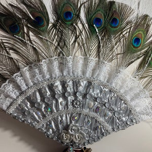 Peut inclure: Un &eacute;ventail argent&eacute; et blanc avec une bordure en dentelle et des plumes de paon. L'&eacute;ventail est d&eacute;cor&eacute; de perles et de strass.