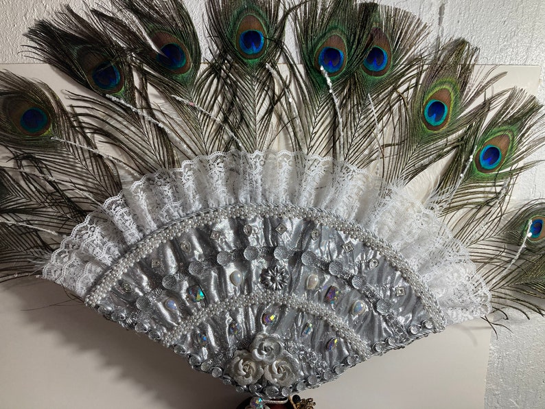 Peut inclure: Un &eacute;ventail argent&eacute; et blanc avec une bordure en dentelle et des plumes de paon. L'&eacute;ventail est d&eacute;cor&eacute; de perles et de strass.
