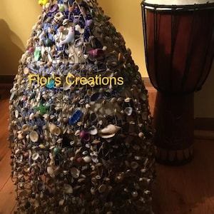 Peut inclure: Un vase en céramique bleu et blanc recouvert d'un filet de ficelle et décoré de coquillages, de perles et d'autres petites babioles. Le vase est posé sur un sol en bois à côté d'un tambour en bois brun. Le texte "Flor's Creations" est visible sur le vase.