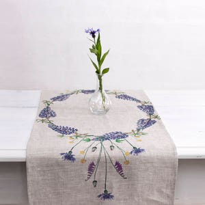 Könnte beinhalten: Ein Tischläufer aus Leinen mit einem Blumendesign, das einen Kranz aus lila Blüten und grünen Blättern zeigt. Eine einzelne blaue Blume steht in einer klaren Glasvase in der Mitte des Läufers.