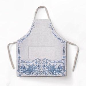 Puede incluir: Un delantal de lino blanco con un patrón de toile azul con diseño floral y un bolsillo. El delantal tiene tirantes ajustables.