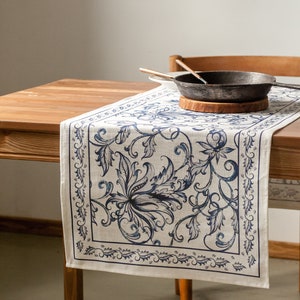 May include: A white linen table runner with a blue floral pattern. The runner is on a wooden table with a cast iron skillet and wooden spoons.