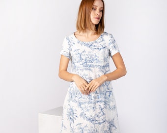 May include: A white linen dress with a blue and white toile pattern. The dress has a scoop neck, short sleeves, and a loose fit. The model is wearing white heeled sandals.