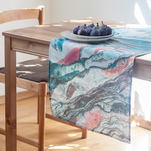 May include: A teal, pink, and brown abstract patterned table runner on a wooden table with a plate of figs.