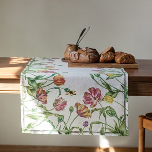 Könnte beinhalten: Ein weißer Tischläufer aus Leinen mit einem Blumenmuster aus Tulpen in Rosa-, Gelb- und Grüntönen. Der Läufer liegt auf einem Holztisch mit Brot auf einem Schneidebrett.