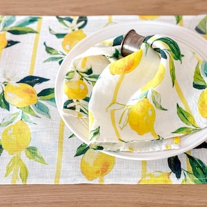 Peut inclure: Une serviette en lin blanche avec un motif imprimé de citron est pliée et placée sur une assiette blanche. La serviette est maintenue en place par un anneau de serviette en argent. L'assiette est sur un set de table rayé jaune et blanc avec un motif imprimé de citron.