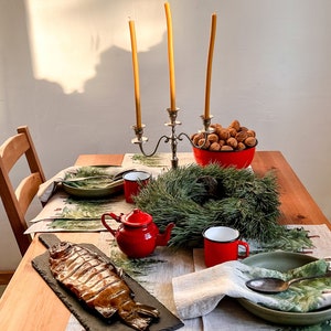 May include: A table setting with a smoked fish on a black slate platter, a red teapot, two red mugs, a green bowl, a green wreath, and a bowl of walnuts. The table is covered in a linen tablecloth with a green and white pattern.