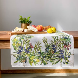 Pode incluir: Um corredor de mesa de linho branco com um design de ramo de oliveira verde. O corredor está sobre uma mesa de madeira com uma pequena planta em vaso, frutas e pastelaria.