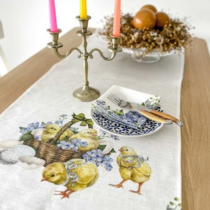 Può includere: Un runner da tavolo in lino bianco con un design floreale con pulcini gialli e fiori blu nontiscordardime. Il runner è decorato con un cestino di pulcini e un piatto con motivo blu e bianco con una forchetta e un coltello.