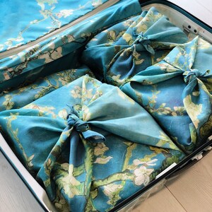 K&ouml;nnte beinhalten: Ein blauer Koffer mit einem floralen Muster als Innenfutter. Der Koffer ist ge&ouml;ffnet und enth&auml;lt vier Gegenst&auml;nde, die in blaues Stoff mit einem floralen Muster gewickelt sind. Der Stoff ist mit Knoten gebunden.