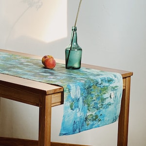 Peut inclure: Une table en bois avec un chemin de table bleu et vert orné d'un motif de nénuphar. Une bouteille en verre vert avec une fleur séchée est posée sur le chemin de table, à côté de deux pêches.