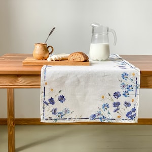 Könnte beinhalten: Ein weißer Tischläufer aus Leinen mit einem blauen Blumenmuster. Der Läufer liegt auf einem Holztisch mit einer Milchkanne, einem kleinen Keramik-Kännchen, einem Messer und einem Stück Brot.