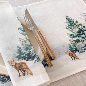 May include: A white linen placemat with a winter forest scene featuring green pine trees and a red fox. The placemat is set with a white plate and a silver fork and knife with wooden handles.