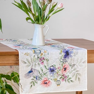 Könnte beinhalten: Weißer Tischläufer mit floralem Aquarelldesign mit rosa, lila und blauen Blumen und grünen Blättern. Eine weiße Kanne mit Tulpen und Samenkapseln steht auf dem Holztisch.