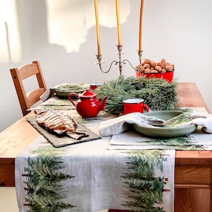 May include: A wooden table set for a festive meal with a red teapot, a plate of smoked fish, and a green bowl with a spoon. The table is decorated with a linen runner featuring a green pine tree design. There are two wooden chairs and a silver candelabra with yellow candles.