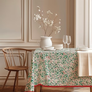 May include: A rectangular tablecloth featuring a repeating pattern of green holly leaves and red berries on a cream background, finished with a solid red border. The fabric drapes over a table set with a white pitcher, bowls, and two tall glasses.