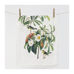 Könnte beinhalten: Ein Leinentuch mit einer botanischen Illustration eines blühenden Baumes mit weißen Blüten, grünen Blättern und gelben Vögeln. Das Tuch hängt an einer Holzklammer an einer Schnur vor weißem Hintergrund.