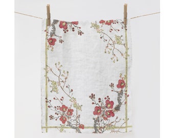 Puede incluir: Un paño de cocina de lino blanco con un diseño floral con flores rojas y amarillas y hojas verdes. La toalla tiene un borde dorado.