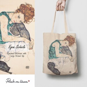 Op de afbeelding: Een tote bag met een schilderij van Egon Schiele getiteld "Zittende vrouw met opgetrokken benen". Het schilderij toont een vrouw met rood haar die zit met haar benen opgetrokken, gekleed in een groene tanktop en zwarte kousen.
