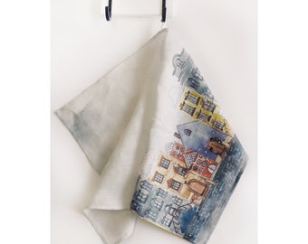 May include: A white linen tea towel with a watercolor illustration of a snowy cityscape. The buildings are painted in shades of blue, yellow, red, and brown. The illustration is surrounded by a blue and white watercolor background.