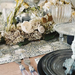 May include: A table setting with a floral centerpiece, a black and white patterned table runner, and a black plate with a silver rim. The centerpiece features white and cream flowers, green leaves, and brown branches. The table runner has a floral design with a black background. The plate is on a wooden table.