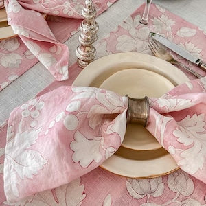 May include: Pink floral linen napkins with white embroidery, folded and placed on a pink floral linen placemat with a silver napkin ring. The placemat is on a light grey linen tablecloth.