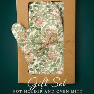 Könnte beinhalten: Ein Geschenkset mit Topflappen und Ofenhandschuh. Die Artikel werden in einer braunen Pappschachtel mit einem Ausschnittfenster präsentiert. Topflappen und Ofenhandschuh haben ein Blumenmuster mit grünen Blättern und orangefarbenen Blüten. Die Worte "Gift Set" und "POT HOLDER AND OVEN MITT" sind auf der Box aufgedruckt.