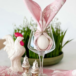 May include: A pink napkin folded into a bunny rabbit shape with a white egg inside, sitting in a clear wine glass. The glass is on a table with a white ceramic rooster and two silver salt and pepper shakers.