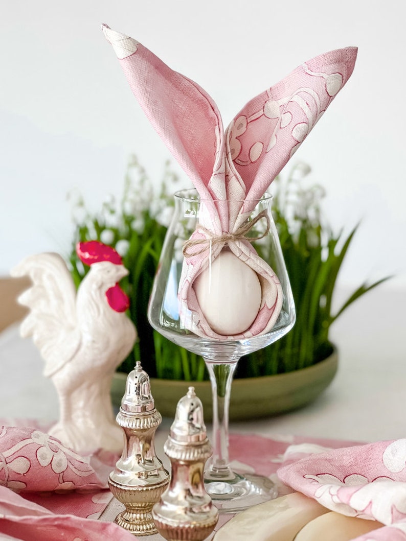 May include: A pink napkin folded into a bunny rabbit shape with a white egg inside, sitting in a clear wine glass. The glass is on a table with a white ceramic rooster and two silver salt and pepper shakers.