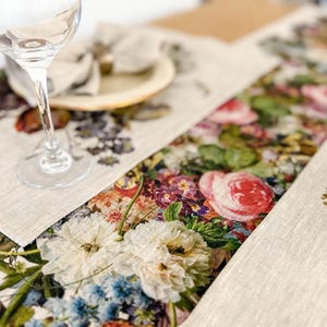 May include: A beige linen table runner with a floral print featuring a variety of colorful flowers, including roses, peonies, and lilies. The runner is placed on a wooden table with a glass of water and a napkin.