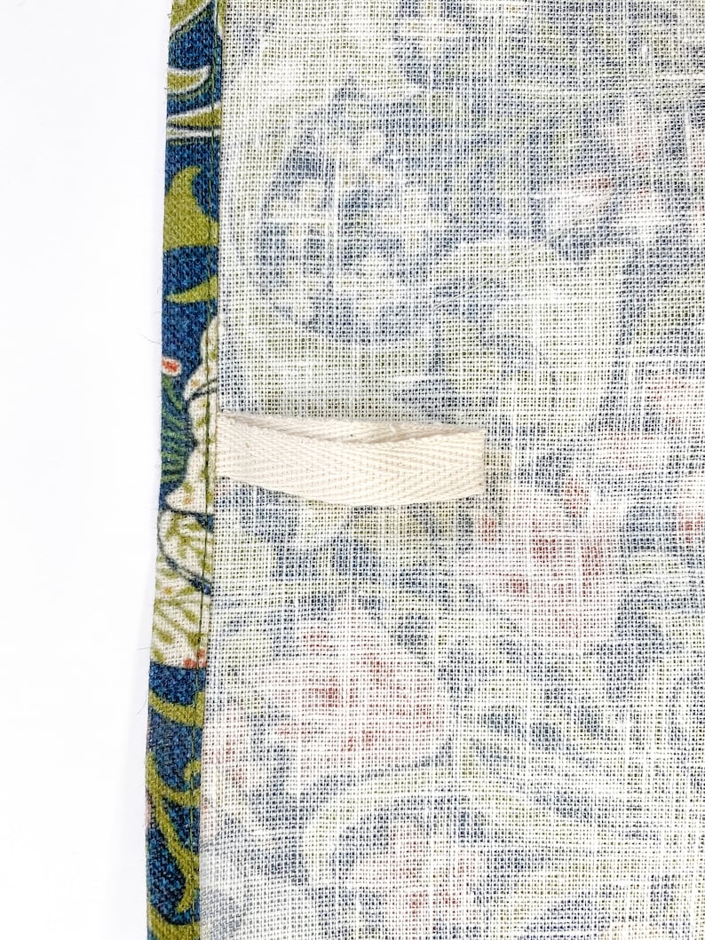 Pu&ograve; includere: Un primo piano di un asciugamano da cucina in lino bianco con un motivo floreale blu e verde. L'asciugamano ha un anello di tessuto color crema cucito sul lato.