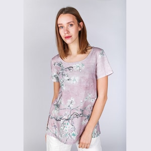 May include: A pale pink linen t-shirt with a floral print of a blooming tree branch. The print is in shades of white, green, and pink.
