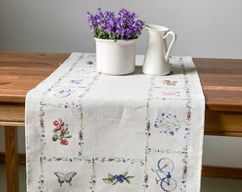 May include: A white linen table runner with a floral design featuring butterflies, bicycles, berries, and flowers. The runner is on a wooden table with a white pitcher and a white pot with purple flowers.
