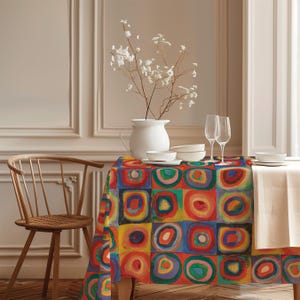Pode incluir: Uma toalha de mesa colorida com um design geométrico de quadrados e círculos concêntricos em vermelho, amarelo, azul e verde. A mesa está posta com louça branca, taças de vinho e um vaso branco com flores.