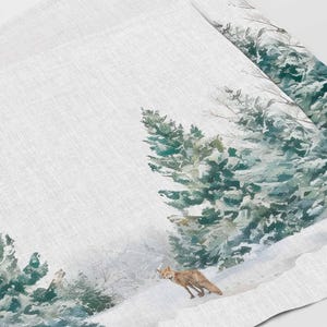May include: A white linen tea towel with a watercolor design of a snowy forest scene. The design features green pine trees, a red fox, and a white snowy background.
