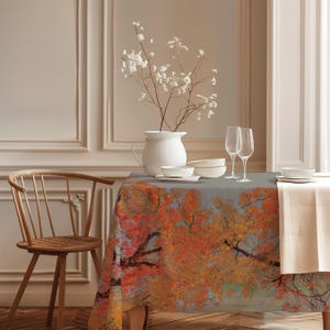 May include: A rectangular tablecloth featuring a vibrant autumn tree print with orange, red, and yellow leaves against a grey background. The table is set with a white ceramic pitcher containing white flowers, two clear glass flutes, and white bowls.