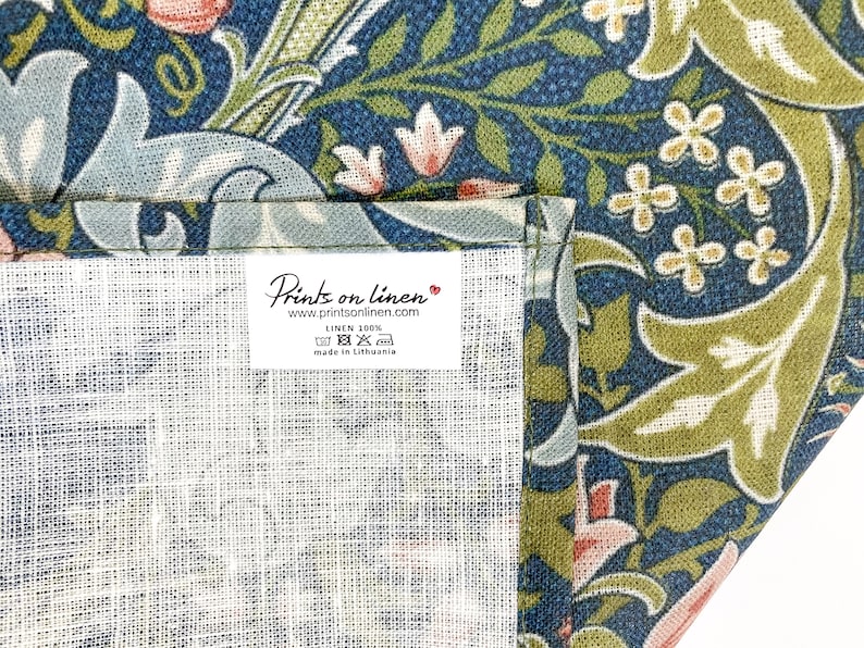 Pu&ograve; includere: Un primo piano di un tessuto di lino bianco con un'etichetta che recita "Prints on linen" e "www.printsonlinen.com". L'etichetta indica anche "Linen 100%" e "Made in Lithuania". Il tessuto presenta un motivo floreale in tonalit&agrave; blu, verde e rosa.