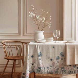 Pode incluir: Uma toalha de mesa de linho bege com uma estampa floral em aquarela com flores rosa, azuis e verdes. A toalha de mesa está posta sobre uma mesa de madeira com um jarro branco, tigelas e taças de vinho.
