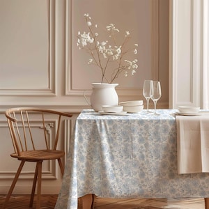 Puede incluir: Un mantel blanco con un patrón floral azul, preparado para una cena con dos copas de vino, platos y un jarrón de flores blancas.