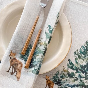 May include: A white linen dinner napkin with a watercolor design of a snowy forest and foxes. The napkin is folded and placed on a white plate with a gold rim. A fork and knife are placed on top of the napkin.