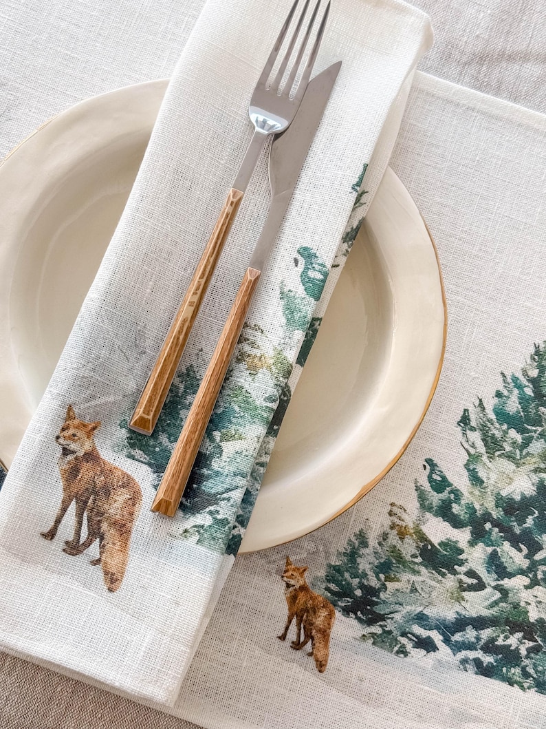 May include: A white linen dinner napkin with a watercolor design of a snowy forest and foxes. The napkin is folded and placed on a white plate with a gold rim. A fork and knife are placed on top of the napkin.