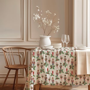 Pode incluir: Uma toalha de mesa retangular com um padrão repetitivo de figuras de quebra-nozes, bengalas doces e folhas de azevinho verdes com bagas vermelhas sobre um fundo creme. O tecido cobre uma mesa posta com tigelas e copos brancos.