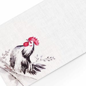 May include: A white linen placemat with a watercolor illustration of a black and white rooster with a red comb and wattle. The rooster is perched on a branch with gray and black leaves.