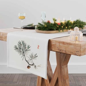 May include: White table runner featuring a pine branch design in shades of green and brown. The runner is displayed on a wooden table with a white lantern and a garland of greenery with lights.