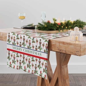 May include: White table runner with a repeating pattern of nutcracker figurines, candy canes, holly, and candles. The runner has a red and blue stripe with the text "Merry Christmas" in green.