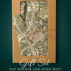 Op de afbeelding: Een cadeau set met een pannenlap en ovenwant met een bloemenpatroon in groen, crème en grijs. De set wordt gepresenteerd in een bruine kartonnen doos met een venster, vastgebonden met touw. Tekst op de doos luidt "Gift Set POT HOLDER AND OVEN MITT."